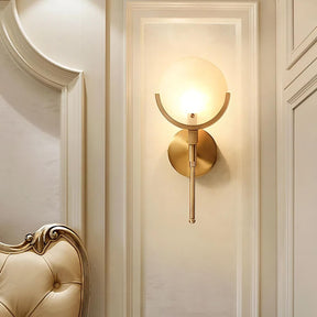 The Moonshade Marble Wall Light Sconce by Morsale.com showcases its sophisticated design with a round, frosted glass shade and a gold metallic fixture, beautifully enhancing an ornate cream-colored wall. A cream tufted headboard is partially visible, adding elegance to the room.