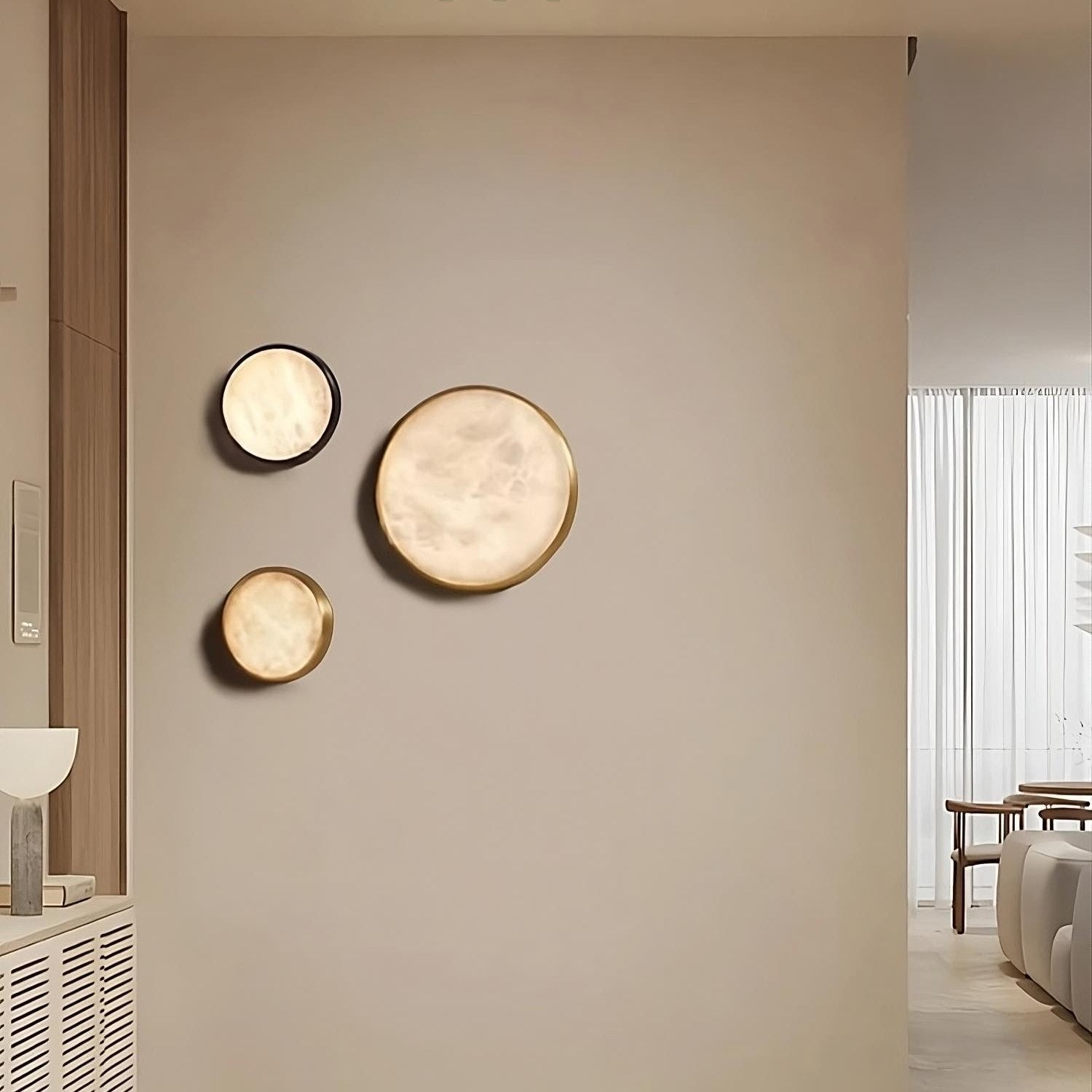 The modern interior showcases Morsale's Moonshade Marble & Copper Light Fixtures, where three circular mirrors of various sizes adorn a neutral-colored wall. The room boasts a minimalist copper design with soft lighting, and you can partially see a white lamp along with a glimpse of a table and chairs in the background.