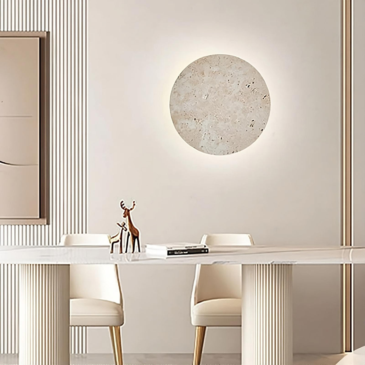 A modern dining room features a sleek white table with two white chairs. Above the table, a round textured wall art piece is softly illuminated by Natural Travertine Wall Sconces from Morsale.com mounted on a beige wall. A minimalist deer figurine and an open book are placed on the table, adding to the serene ambiance.