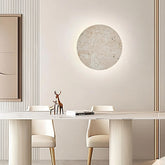 A modern dining room features a sleek white table with two white chairs. Above the table, a round textured wall art piece is softly illuminated by Natural Travertine Wall Sconces from Morsale.com mounted on a beige wall. A minimalist deer figurine and an open book are placed on the table, adding to the serene ambiance.