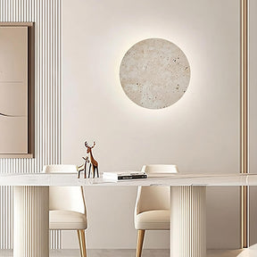 A modern dining room features a sleek white table with two white chairs. Above the table, a round textured wall art piece is softly illuminated by Natural Travertine Wall Sconces from Morsale.com mounted on a beige wall. A minimalist deer figurine and an open book are placed on the table, adding to the serene ambiance.