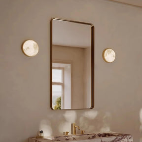 A modern bathroom showcasing a large rectangular mirror with a sleek metal frame, flanked by two round lights from the Moonshade collection by Morsale. Below is a luxurious marble countertop featuring gold fixtures. The space is softly illuminated by copper light fixtures, while a window in the background allows natural light to filter in, creating a serene atmosphere.