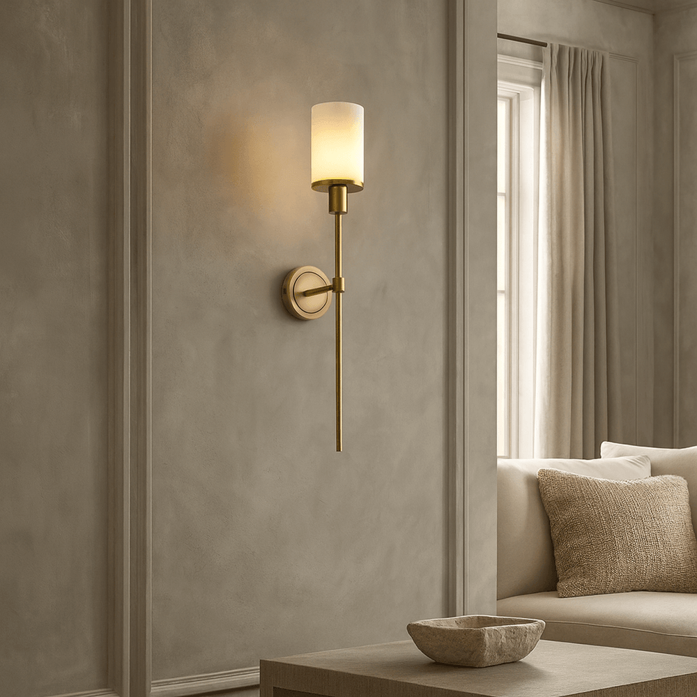 The Morsale Caldera Marble & Copper Wall Sconce with a frosted glass shade is mounted on a gray paneled wall near a window with beige curtains, with a beige sofa and a natural marble bowl on the table in the foreground.