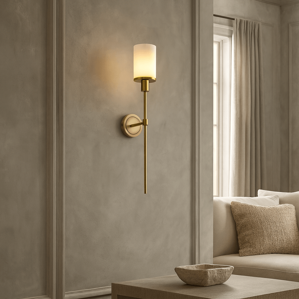 The Morsale Caldera Marble & Copper Wall Sconce with a frosted glass shade is mounted on a gray paneled wall near a window with beige curtains, with a beige sofa and a natural marble bowl on the table in the foreground.