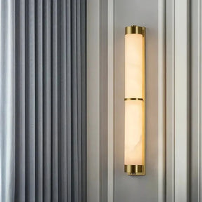 A sleek, modern 23" Natural Marble & Brass Wall Light by Morsale with a brass frame and cylindrical frosted white glass shade is mounted on a light gray paneled wall. Featuring an energy-efficient LED design, this fixture embodies sustainable luxury. On the left side of the image, a floor-to-ceiling gray curtain is partially visible.