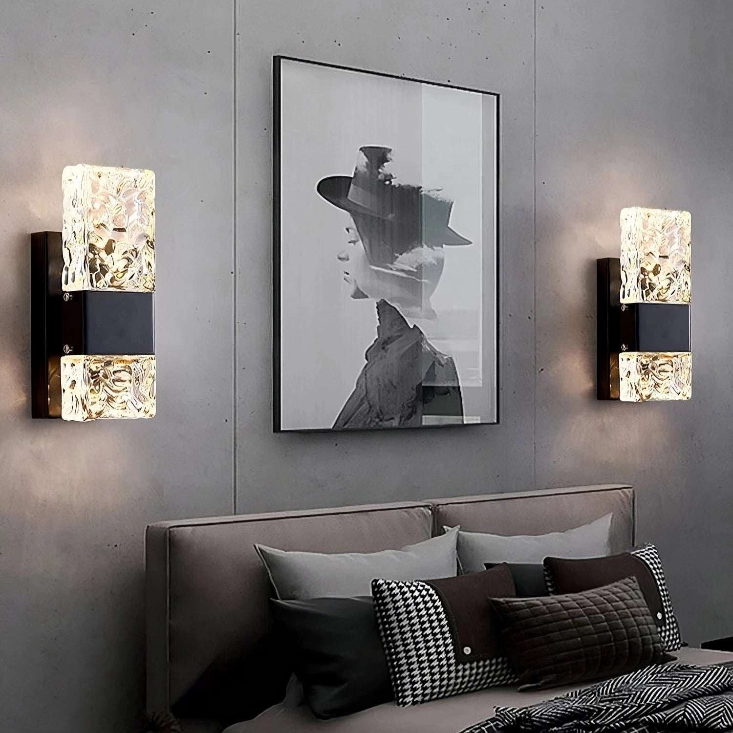 A modern bedroom with a gray wall showcases a black-and-white portrait of a woman in a hat. Two Water Ripple Crystal Wall Sconces by Morsale, featuring textured glass and touch on/off switches, cast warm light. Below, the bed is adorned with several decorative pillows in shades of gray and black.