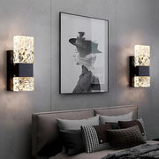 A modern bedroom with a gray wall showcases a black-and-white portrait of a woman in a hat. Two Water Ripple Crystal Wall Sconces by Morsale, featuring textured glass and touch on/off switches, cast warm light. Below, the bed is adorned with several decorative pillows in shades of gray and black.