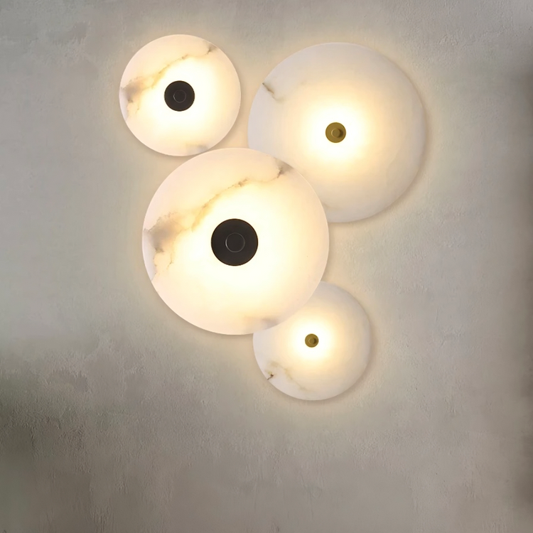 The Moonshade Natural Marble Wall Sconce set by Morsale.com includes four circular lights of varying sizes, emitting a warm glow. Their elegant marble design on a textured wall adds a modern and artistic ambiance.