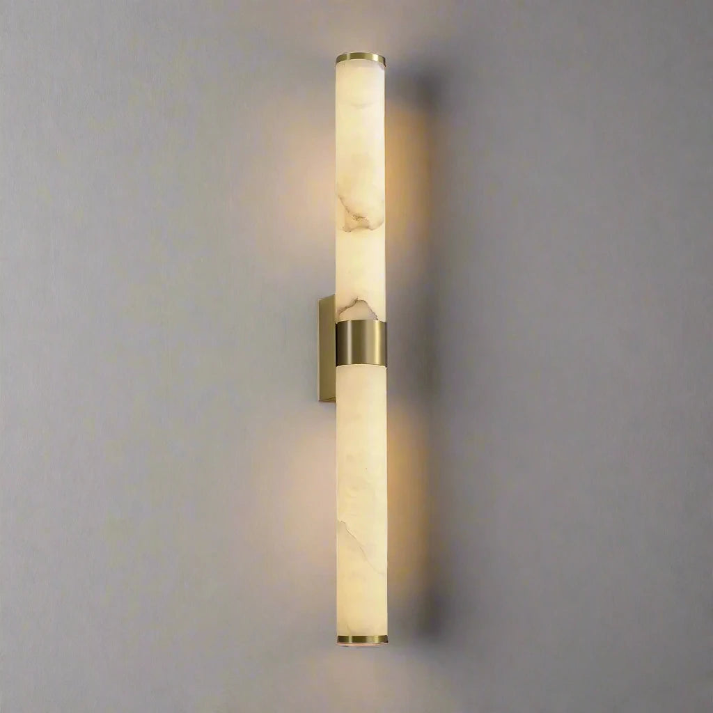 The 27" Moonshade Natural Marble & Copper Wall Sconce by Morsale features a contemporary design with two cylindrical natural marble shades arranged vertically on a copper fixture against a gray wall. It emits a gentle illumination from both ends, creating a soft, ambient glow.