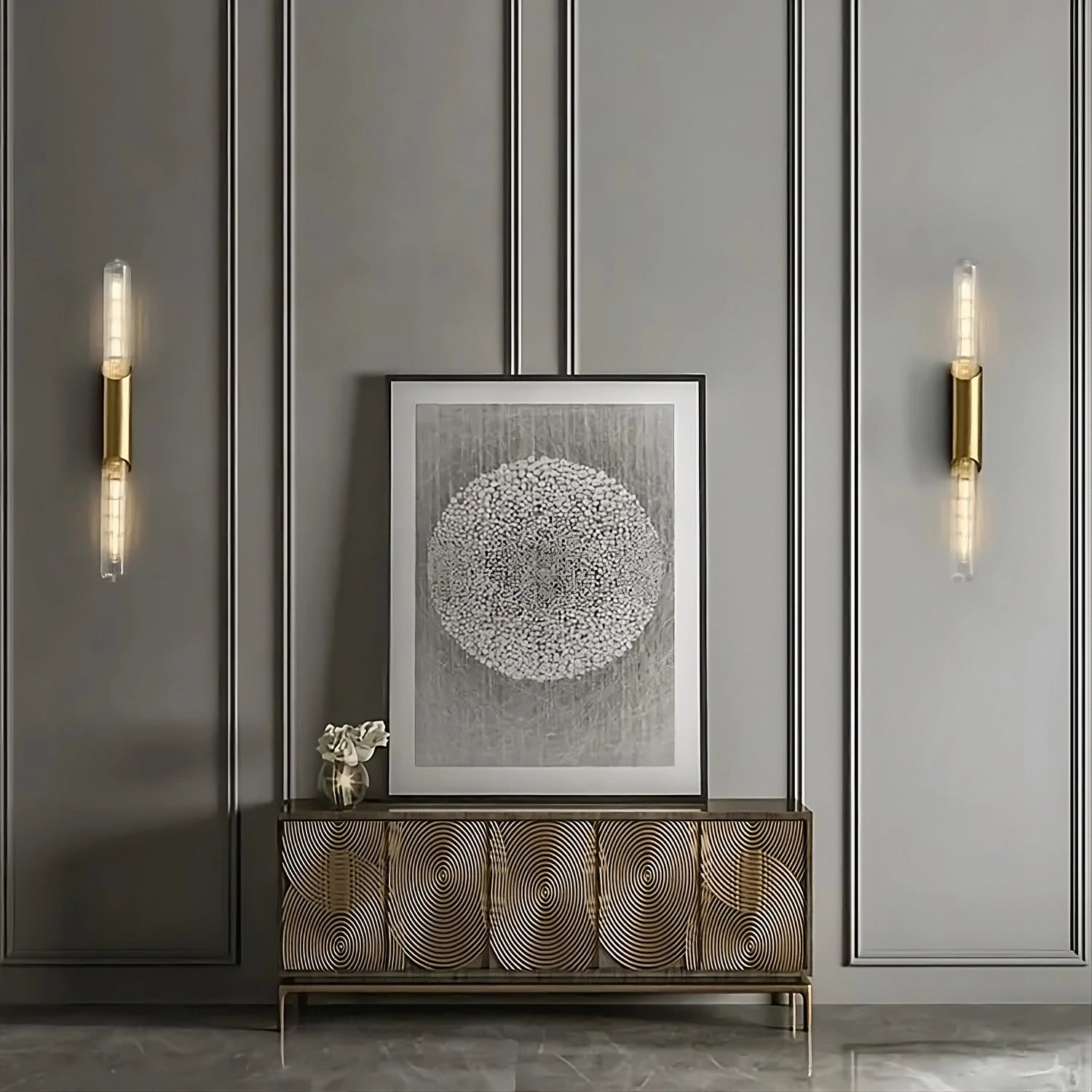 A modern interior wall features vertical paneling in a soft gray color. Centered is a framed abstract artwork with a circular design. Below it stands a wooden sideboard with a decorative, wavy pattern. Two elegant Morsale Palermo 23" Brass Wall Sconce Lights with cylindrical glass fixtures flank the artwork.