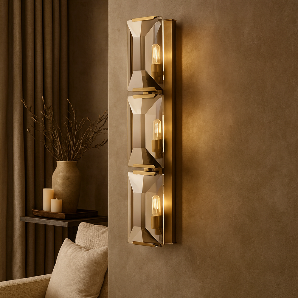 The Brookhill Crystal Wall Sconce by Morsale.com features three geometric glass and brass fixtures, mounted on a taupe wall to softly illuminate a cozy living room with beige furniture, a vase, and candles in the background.