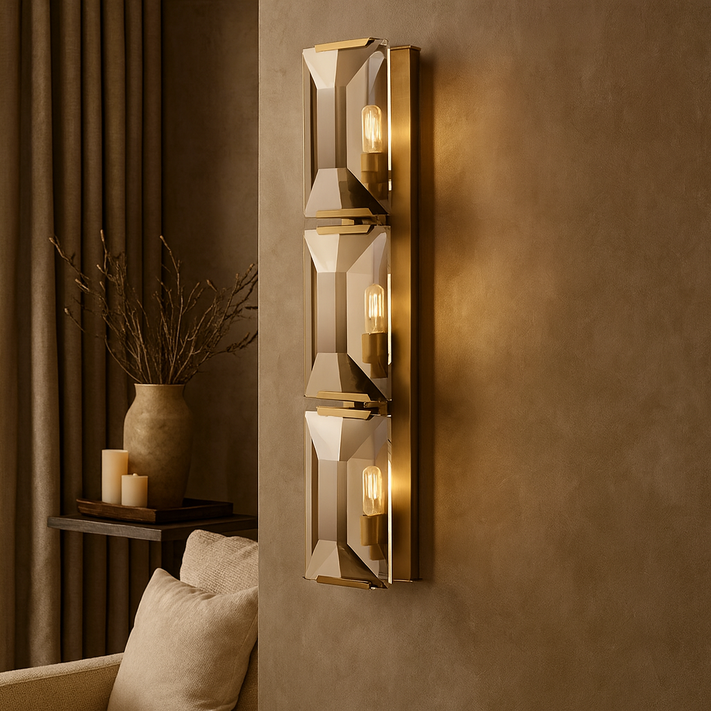 The Brookhill Crystal Wall Sconce by Morsale.com features three geometric glass and brass fixtures, mounted on a taupe wall to softly illuminate a cozy living room with beige furniture, a vase, and candles in the background.