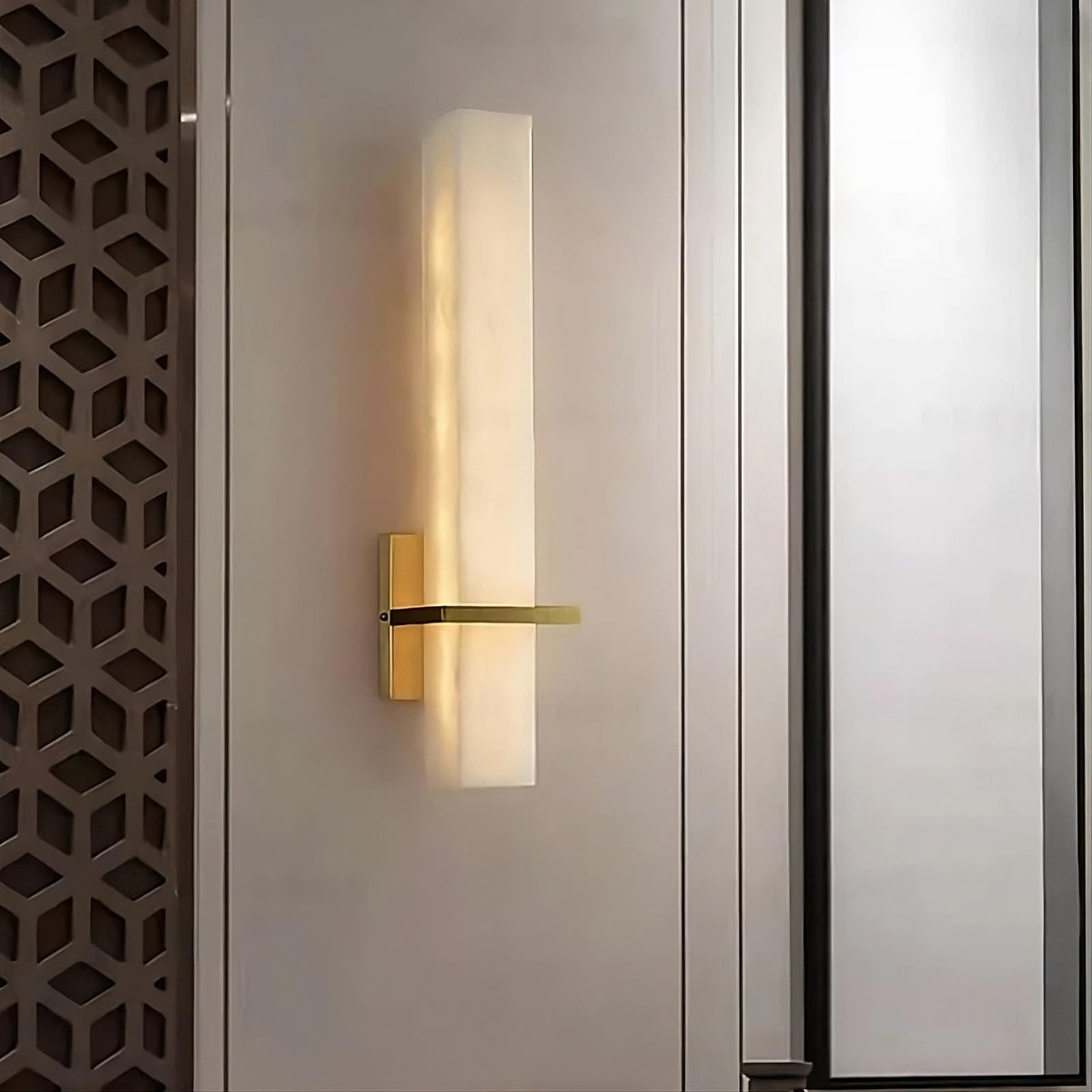 A modern wall light with a rectangular frosted glass shade and a gold-colored base is mounted on the wall. Featuring an elegant design, the wall displays a geometric lattice on the left and a vertical panel on the right. This Natural Marble & Brass Wall Sconce by Morsale.com emits a soft, warm glow throughout the room.