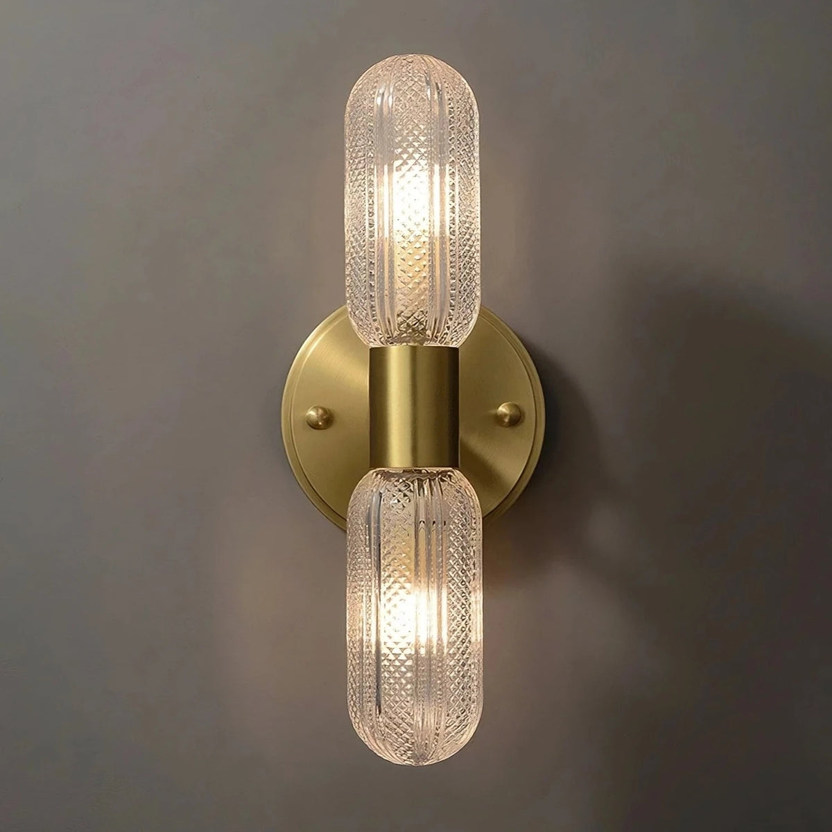 A Morsale Palermo 12" Brass Wall Light Sconce with a sleek base and two vertically aligned, cylindrical, textured glass bulbs that emit a warm glow. With its modern design, this fixture becomes an elegant addition to your home decor. The text "Turn on" is displayed below the fixture.