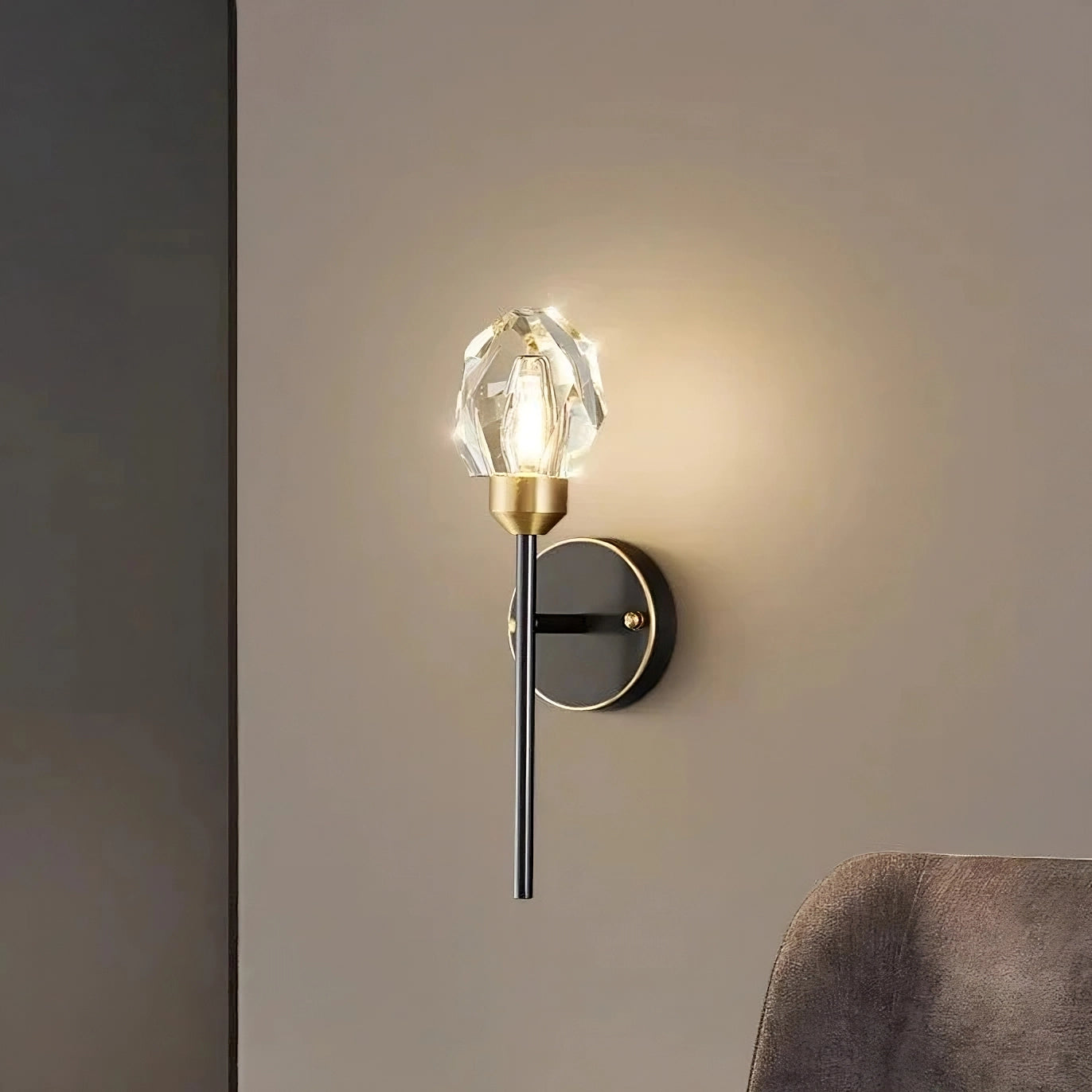 The Morsale Diamante Crystal Wall Sconce, featuring a brass-finished arm and a faceted glass shade, casts a warm glow against the beige wall. In the bottom right corner, part of a gray upholstered chair is visible, enhancing your home decor with elegance and ensuring optimal lighting.