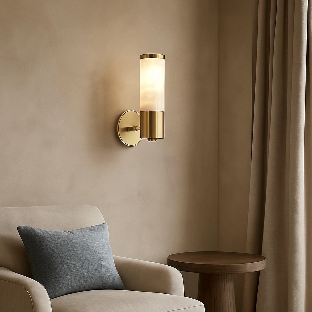 The Moonshade Natural Marble Wall Sconce by Morsale.com, featuring a cylindrical frosted glass shade and brass accents, glows above a beige armchair with a blue-gray pillow, beside a round wooden side table and beige curtains.