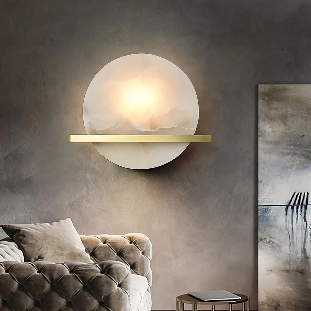A modern, elegant living room features the Natural Marble Wall Light Sconce by Morsale.com with an LED light source, emitting a diffuse glow, and a minimalist golden horizontal bar. Below is a tufted beige sofa, and next to it, a small round side table. A piece of abstract art hangs on the adjacent wall.