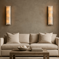 A beige sofa with four large cushions sits against a taupe wall, flanked by two Brookhill Calcite Wall Sconces from Morsale.com. In front, a wooden coffee table holds a decorative bowl and two stacked books.