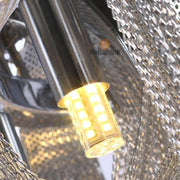 Close-up of a Cadena Chain Tassel Modern Chandelier emitting a warm yellow glow. The chandelier is encased in a sleek silver metallic fixture with a textured mesh design, creating a stylish and contemporary look. An adjustable ceiling wire length complements the setup, with the brand "Morsale.com" faintly visible in the background.