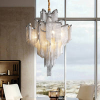 The Cadena Metal Tassel Chandelier by Morsale.com, featuring cascading silver chains and exposed bulbs, hangs above a glass table with two chairs and cups in a modern dining space with large windows overlooking a cityscape.