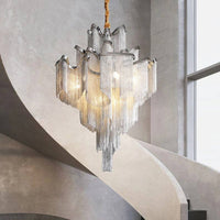 The Cadena Metal Tassel Chandelier by Morsale.com hangs above a curved sculptural staircase in a minimalist space with gray walls and large windows, featuring elegant cascading metal tassels for a modern touch.